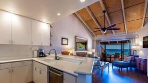 A modern kitchen with white cabinets, a built-in dishwasher, and a large island with a sink. There is a dining area with wooden furniture and a view of the ocean.