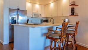 A modern kitchen with white cabinets, a stainless steel refrigerator, a microwave, and a sink. There are two wicker chairs with cushions at the island.