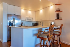 Eine moderne Küche mit weißen Schränken, einem Edelstahlkühlschrank, einer Mikrowelle und einem Spülbecken. An der Kücheninsel stehen zwei Korbstühle mit Kissen.