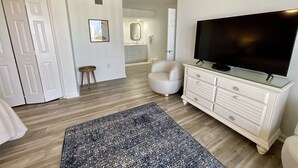 A room with a large flat-screen TV, a white dresser, a patterned rug, and a white chair. - 1703 Brighton, Newly Remodeled Condo With Full Access! 3 Bedroom Condo (Myrtle Beach)