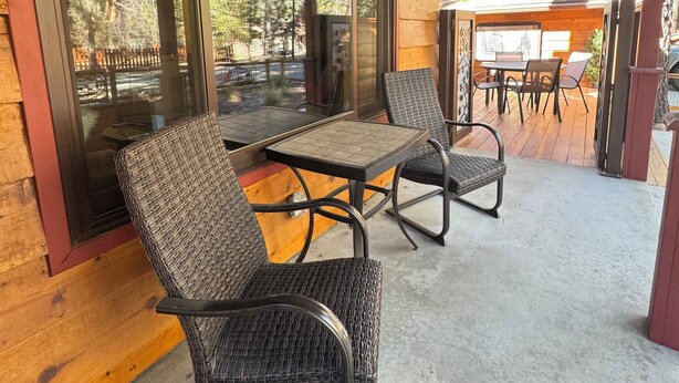 Ein überdachter Patio-Bereich mit Korbstühlen und einem Tisch, großen Fenstern und Blick ins Grüne.