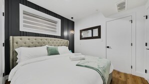 A bedroom with a tufted headboard, a large bed with white linens, a green throw blanket, a bedside lamp, and a window with blinds. - Elegant|Spacious|King Bed|Fireplace|Ravine View (Edmonton)