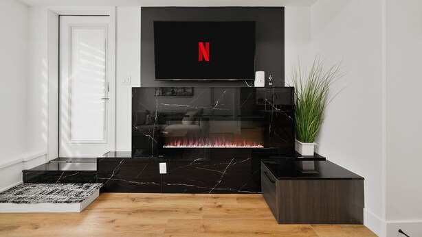 A modern living room with a black marble fireplace, a flat-screen TV mounted above it, and a potted plant on the right. - Elegant|Spacious|King Bed|Fireplace|Ravine View (Edmonton)