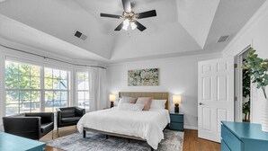 A bedroom with a large bed, two bedside tables, a ceiling fan, and a view of the outdoors through large windows.