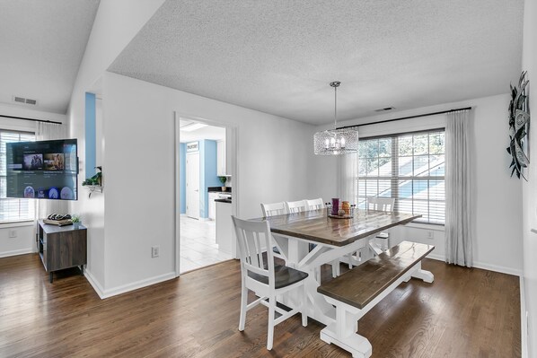 A modern dining area with a wooden table and white chairs, a chandelier, and a view of the outside through large windows.