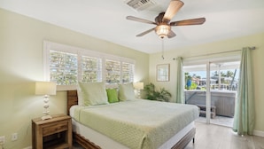 Un dormitorio con cama, mesita de noche, ventilador de techo, ventana con persianas y vista a la zona de la piscina.