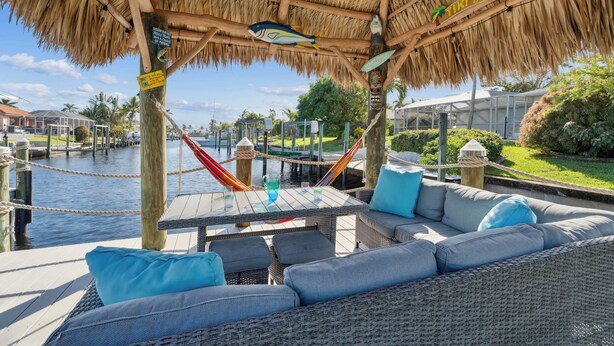 Un bar tiki con techo de paja, asientos exteriores con cojines y vistas a un puerto deportivo.
