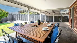 A covered patio with a wooden table set for dining, surrounded by chairs and a brick wall. - Spacious | Pet Friendly | Ping Pong Table | Near Beach (Ocean Grove)