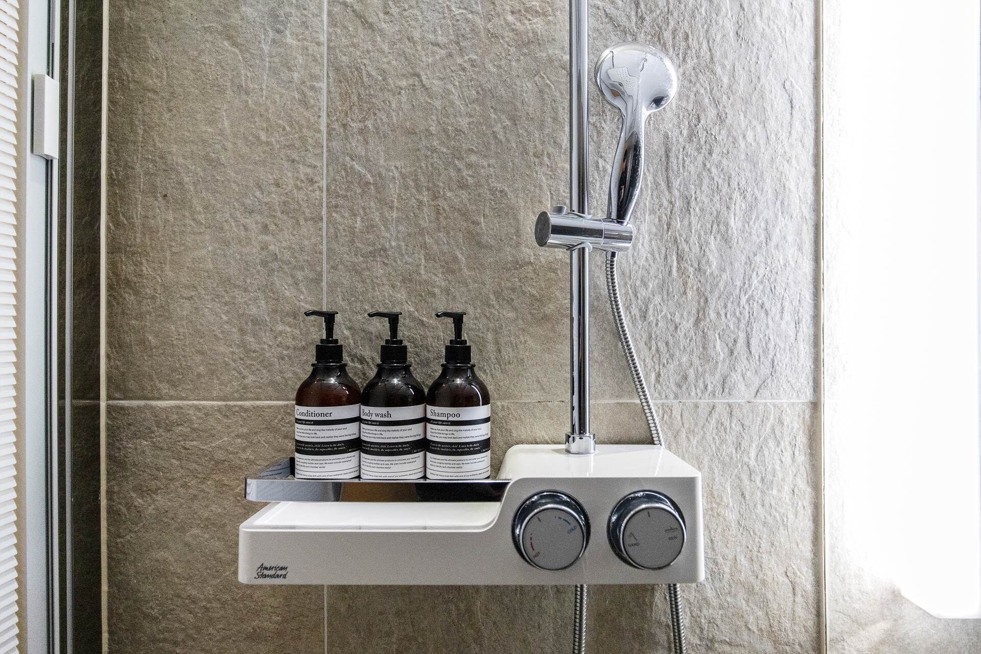 A shower with a handheld showerhead and a wall-mounted control panel. Three bottles of shampoo, conditioner, and body wash are placed on the panel.