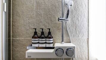 A shower with a handheld showerhead and a wall-mounted control panel. Three bottles of shampoo, conditioner, and body wash are placed on the panel.