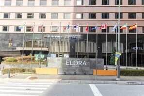 A hotel entrance with multiple flags, a 7-Eleven coffee shop, and a sign for the Elora Hotel. - HOTEL ELORA (Yongin)