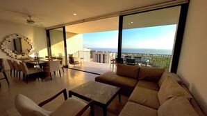 A modern living room with a large sofa, a coffee table, and a dining area with a circular mirror. A balcony with a view of the city is visible through large windows. - Exclusive Solaria Arch View Penthouse with Jacuzzi (Cabo San Lucas)