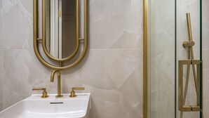 A bathroom with a white sink, gold faucets, a gold-framed mirror, and a glass-enclosed shower with gold fixtures. - GuestReady - Fitzrovia Comfort (London)