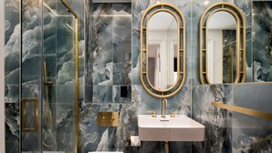 A modern bathroom with a glass-enclosed shower, a white sink with gold fixtures, and marble-patterned walls. - GuestReady - Elegant stay in Fitzrovia (London)