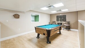 Une table de billard dans une pièce dotée d’un plancher en bois, d’un ventilateur de plafond et d’une fenêtre donnant sur un paysage verdoyant.
