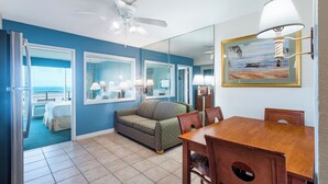 A room with a blue wall, a ceiling fan, a dining table with chairs, a sofa, and a large mirror reflecting a beach scene. - One Bedroom Condo, Oceanside! (Virginia Beach)