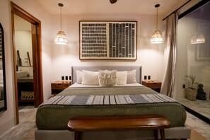 A bedroom with a large bed, two bedside tables, a bench, a wall-mounted artwork, and a large window with curtains.