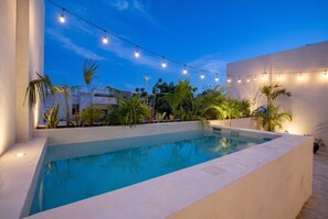 A rectangular swimming pool with a white border, surrounded by greenery and palm trees, under string lights. - Luxury Penthouse w/ Jungle Views (Tulum Municipality)