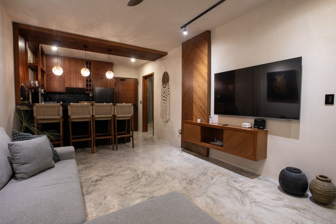 A modern living room with a grey sofa, wooden dining chairs, a TV mounted on a wooden wall, and a kitchen area in the background.