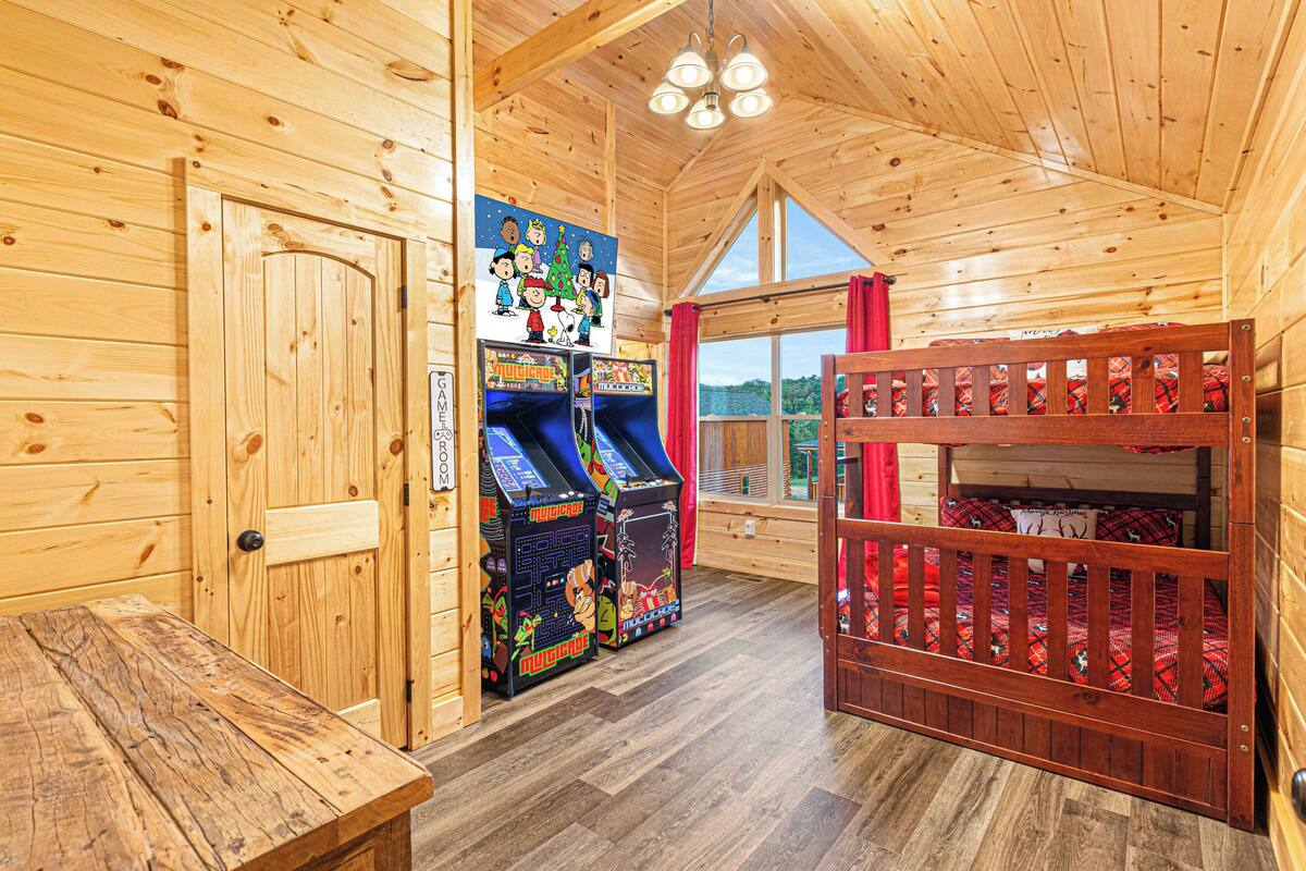 Interior de una cabaña de madera con literas, juegos de arcade y una mesa de madera.