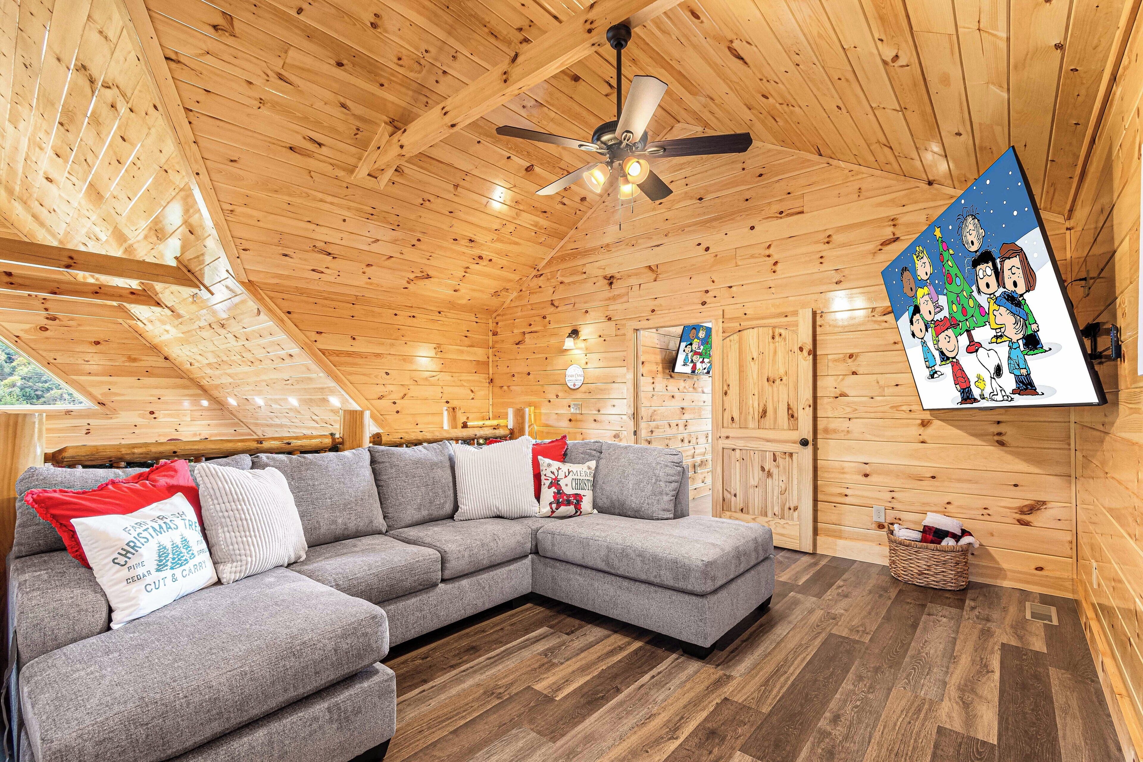 A cozy log cabin interior with a sectional sofa, wooden walls, a ceiling fan, and a wall-mounted TV displaying animated characters.