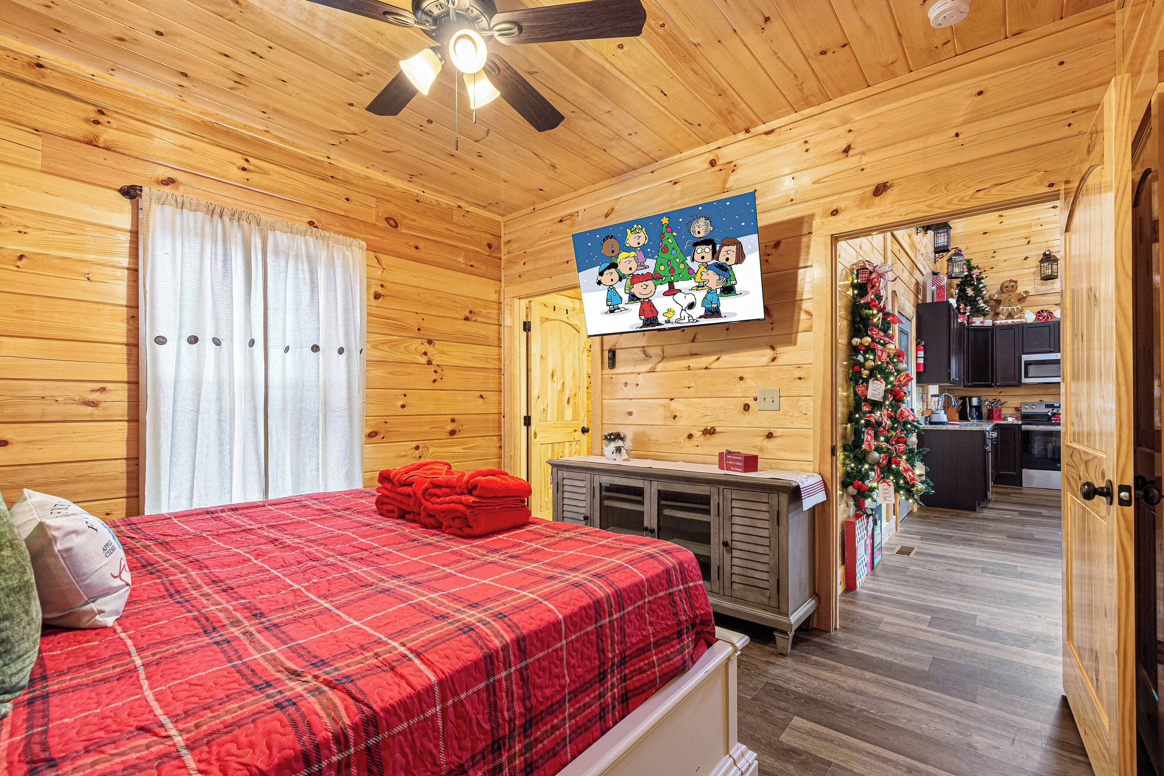 A cozy wooden cabin room with a plaid bedspread, a ceiling fan, a TV mounted on the wall, and a festive Christmas tree in the adjacent kitchen.