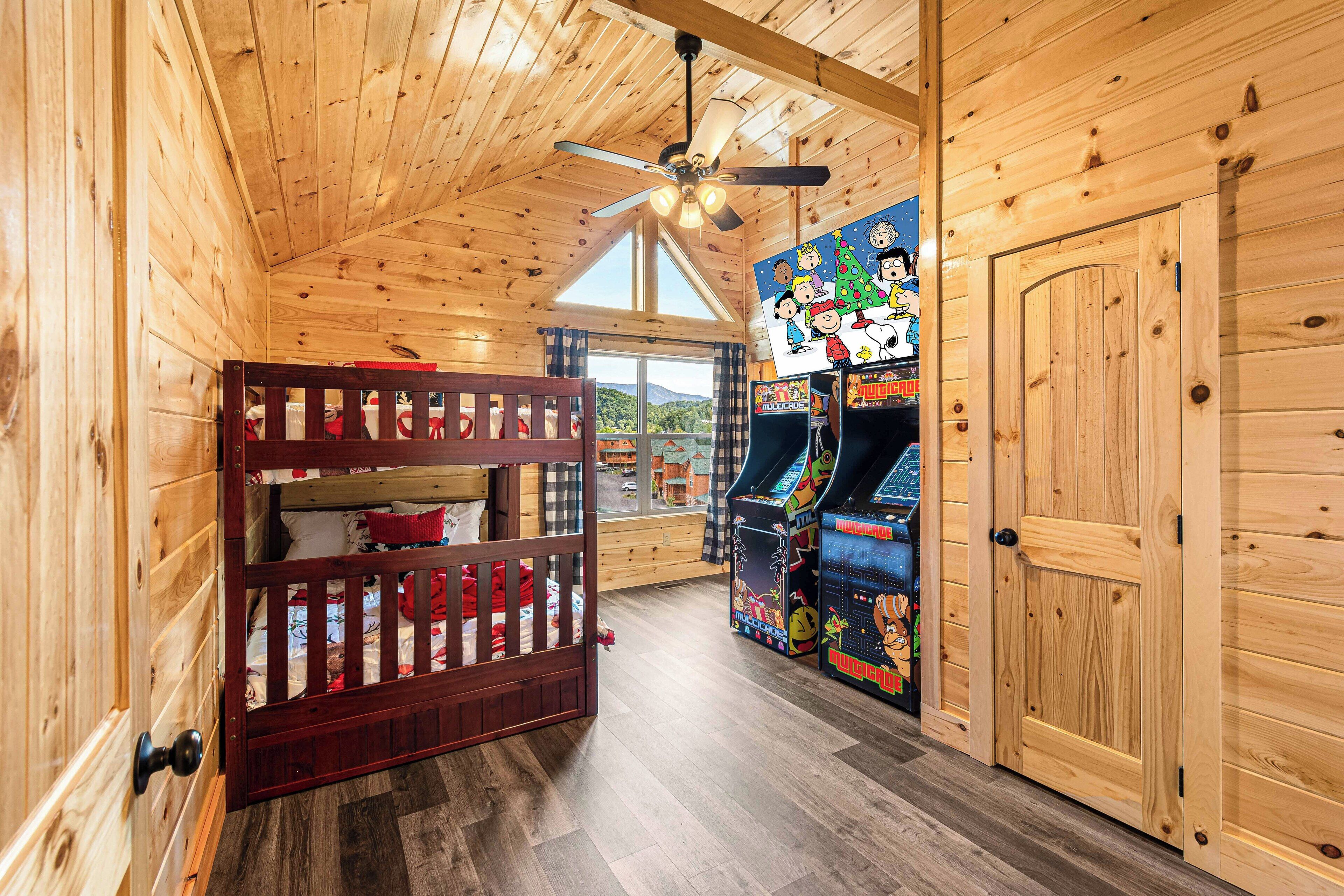 Interior de una cabaña de madera con literas, juegos de arcade y un ventanal grande.