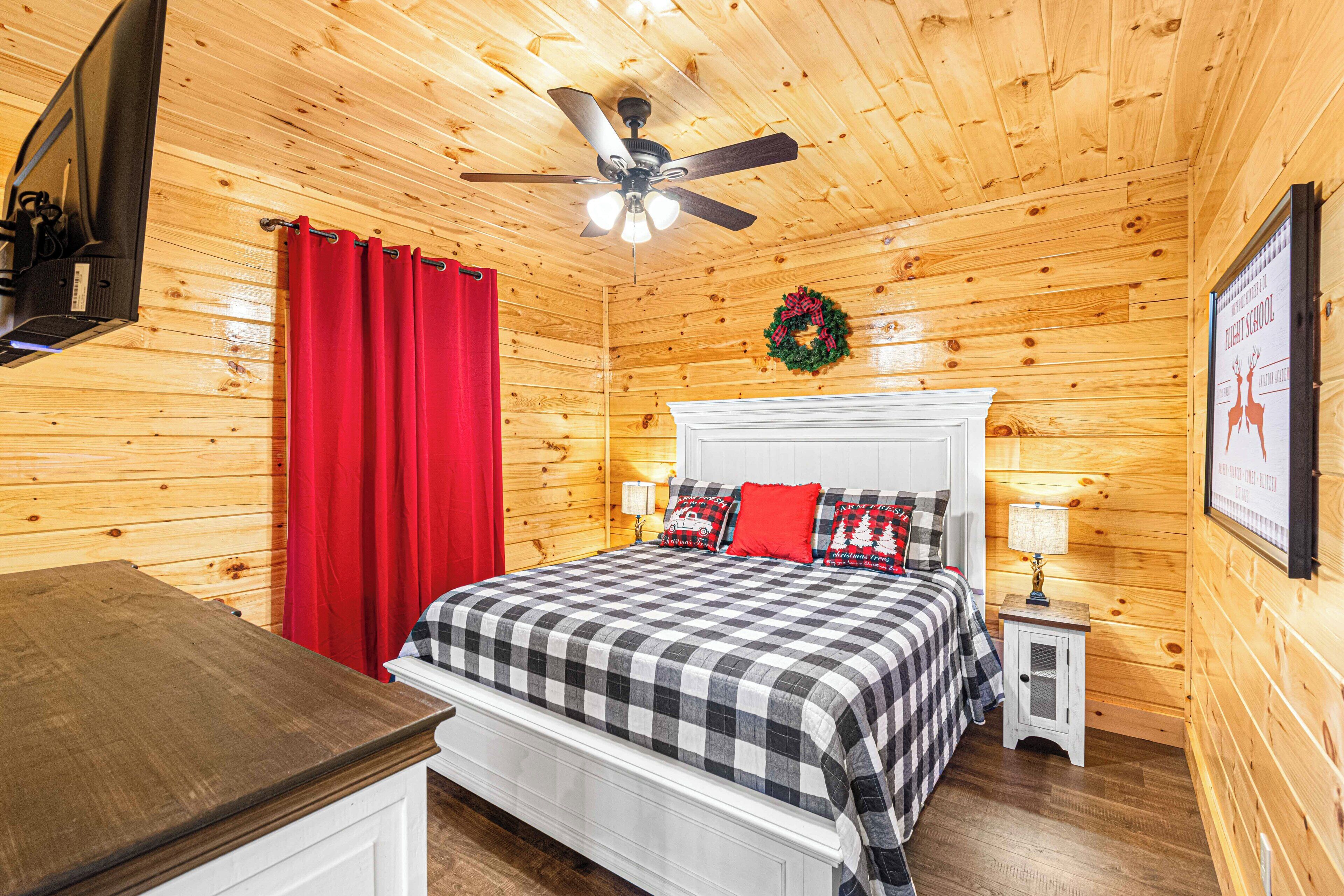 A wooden cabin bedroom with a bed, red curtains, a ceiling fan, a small bedside table, and a framed picture on the wall.