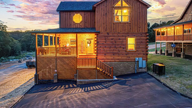 A log cabin with a porch and a covered deck, surrounded by a parking area and trees. - XmasCabins by SmokyStays - Christmas Themed Year Round (Pigeon Forge)