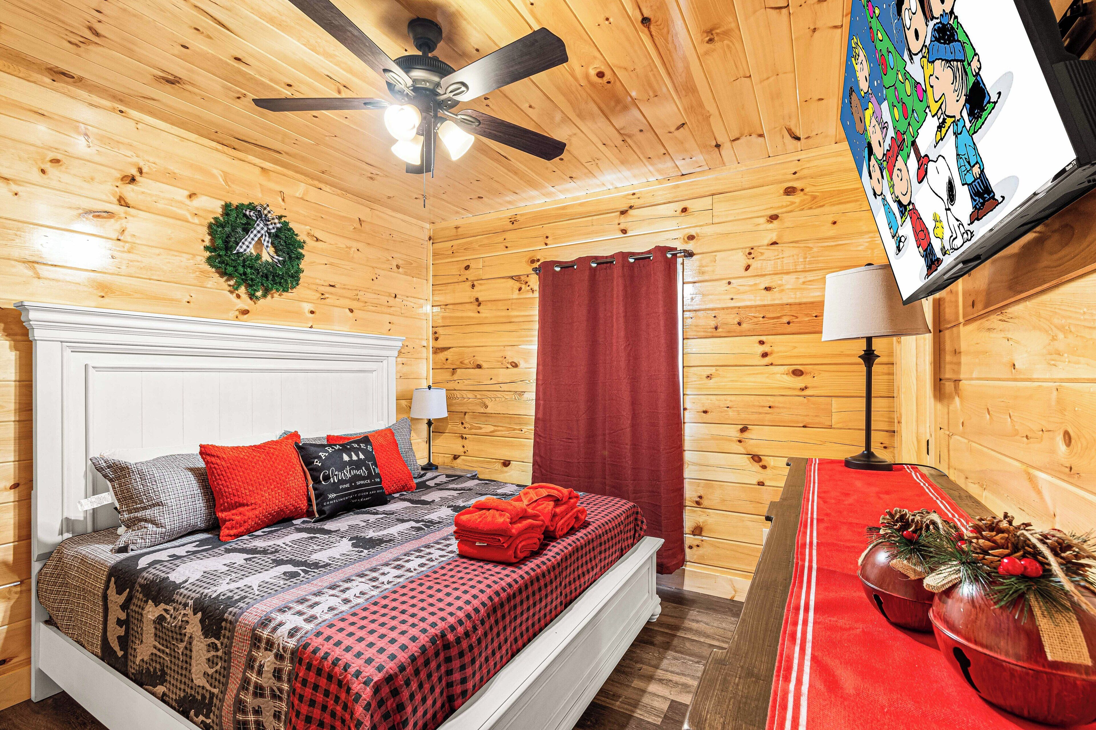A cozy wooden cabin room with a bed, a ceiling fan, a TV, and a Christmas tree decoration.