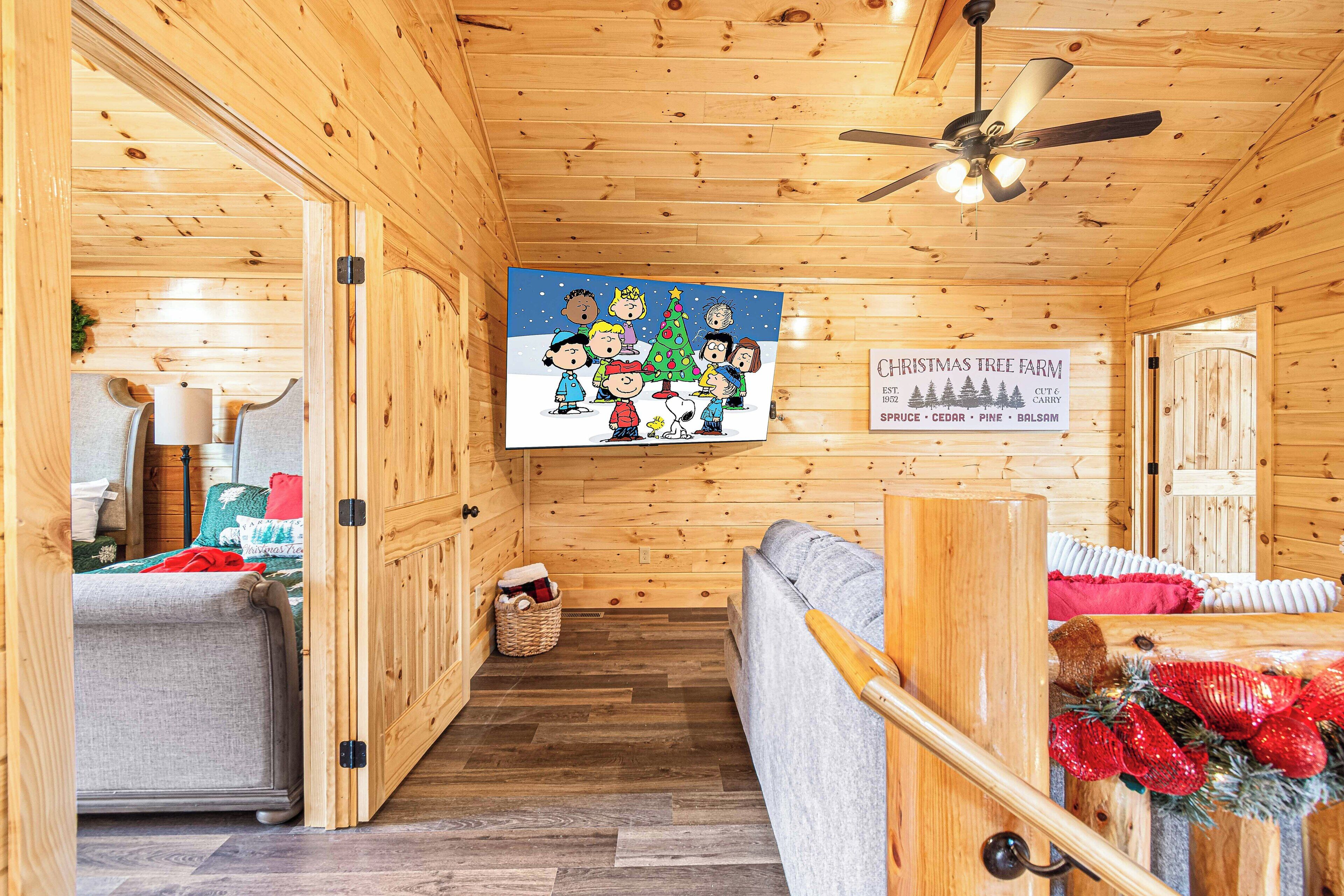 A cozy cabin interior with a wooden ceiling, a ceiling fan, a sofa, a dining table, and a Christmas-themed wall decoration.