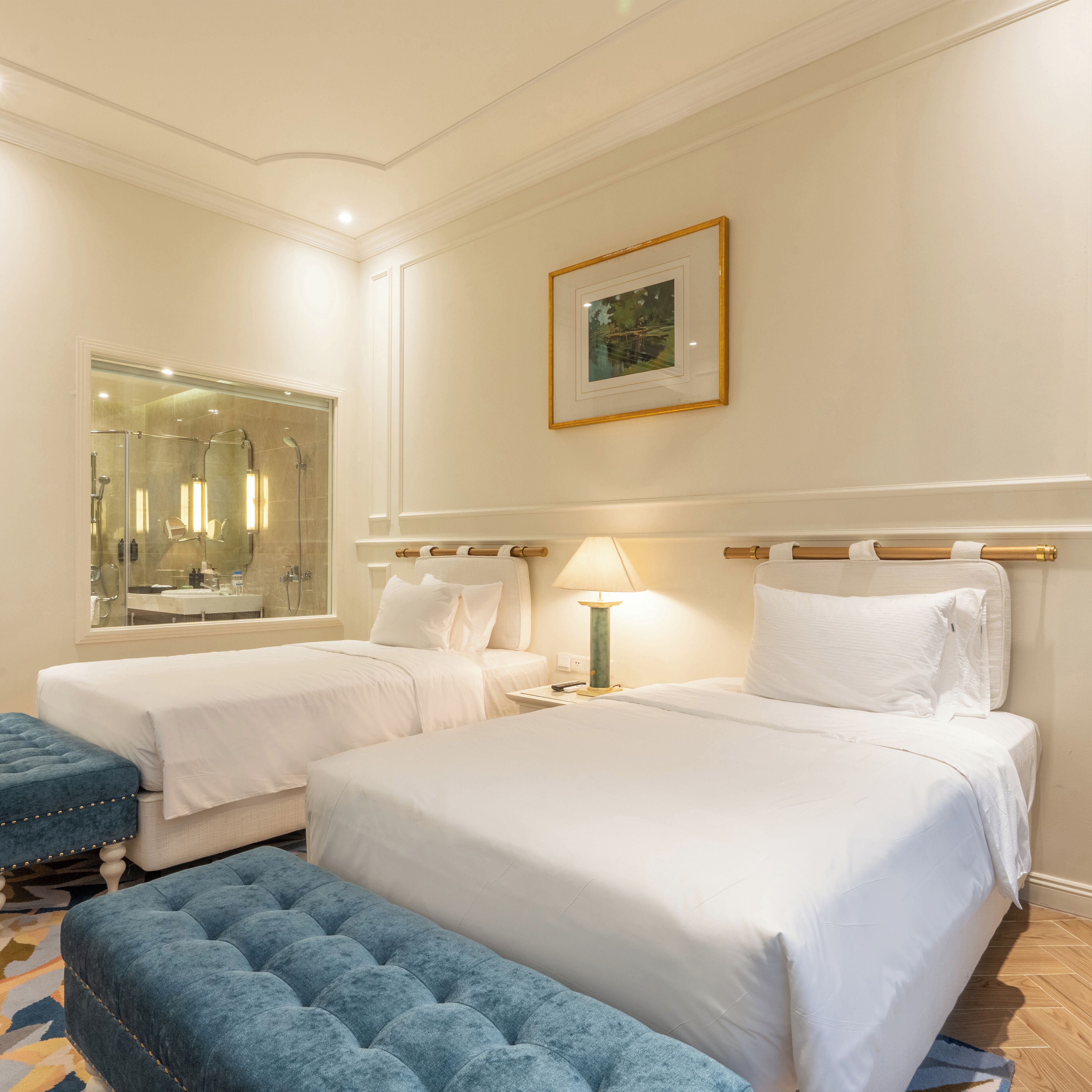 A hotel room with two beds, a large mirror, a framed picture on the wall, and a blue ottoman.