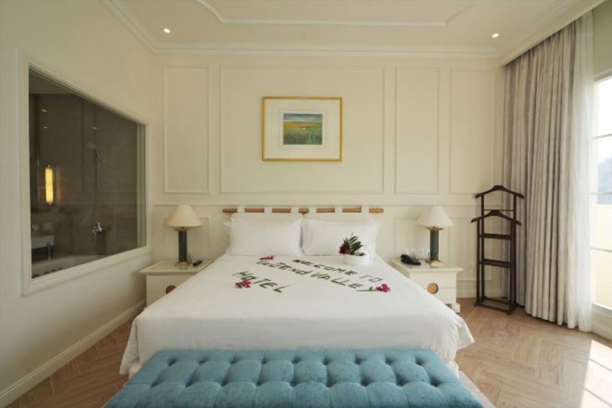 A hotel room with a large bed, a bedside table with lamps, a window with curtains, and a framed picture on the wall.
