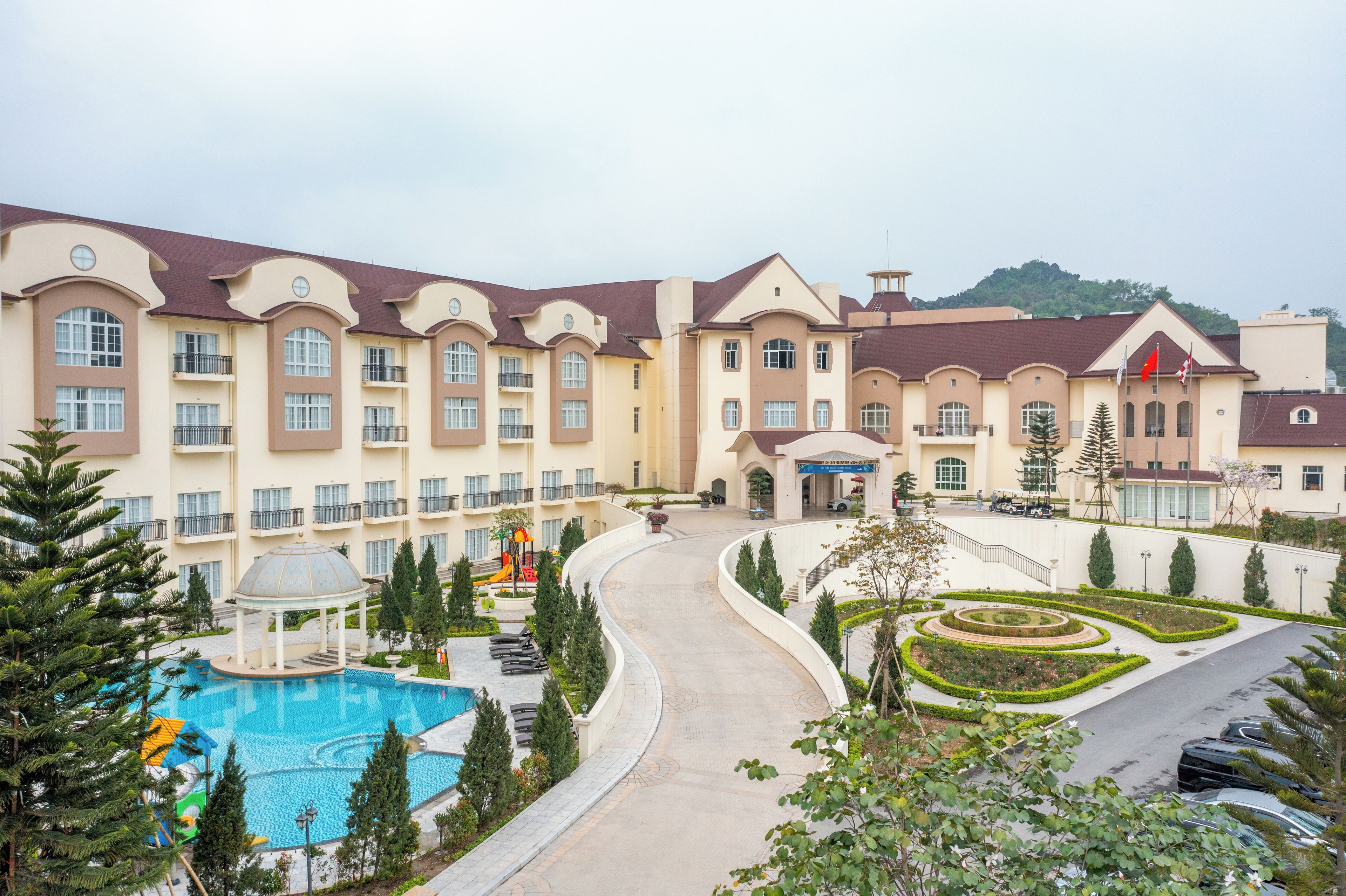 A hotel with a swimming pool, landscaped gardens, and a parking area.