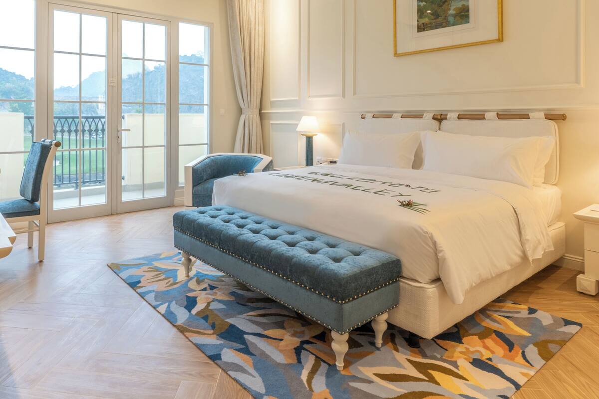 Une chambre avec un grand lit, un banc recouvert d’un tissu bleu, une chaise et une vue sur l’extérieur à travers les portes vitrées.