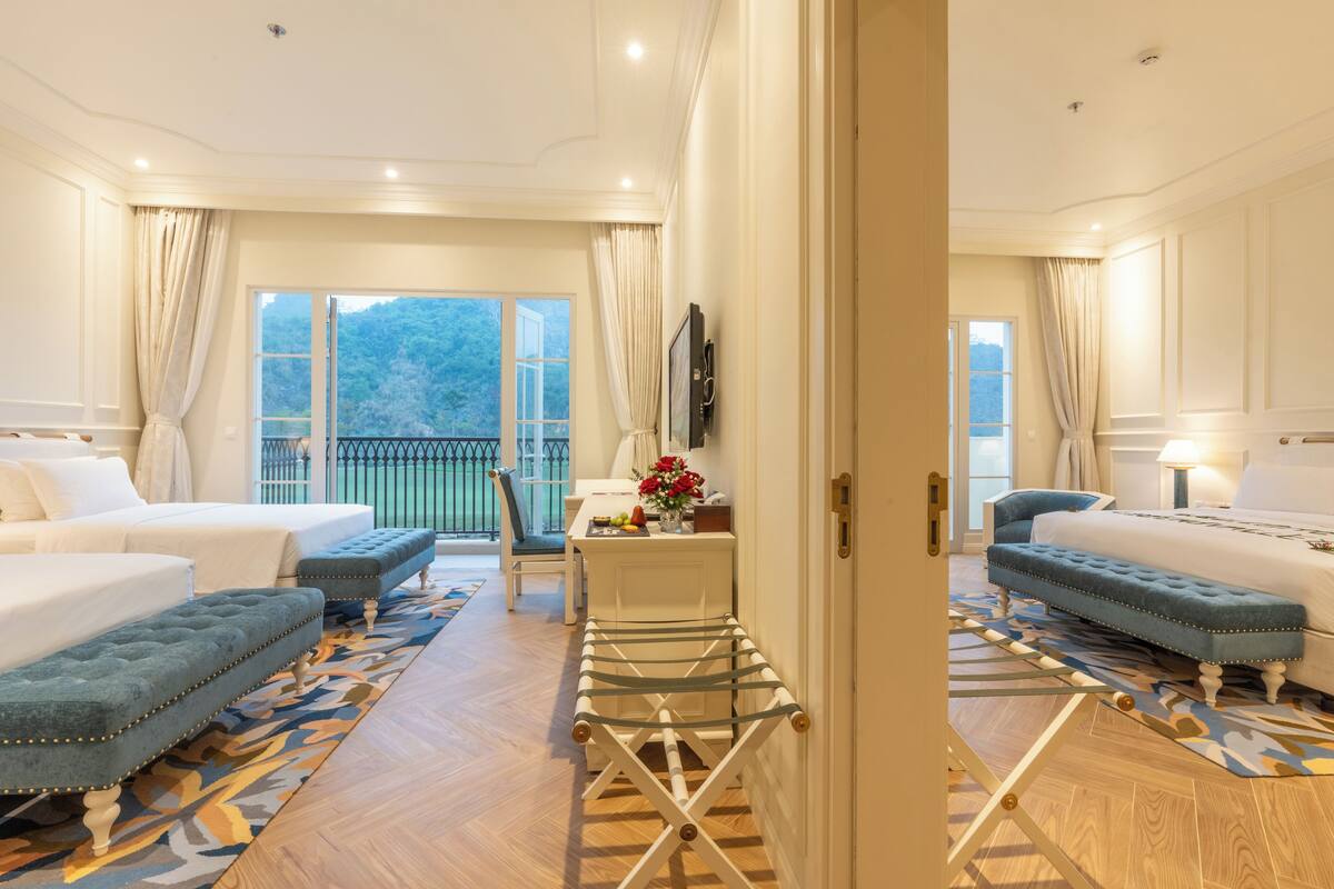 Une chambre d’hôtel avec deux lits, une petite table ornée d’un vase de fleurs et un balcon offrant une vue sur les montagnes.