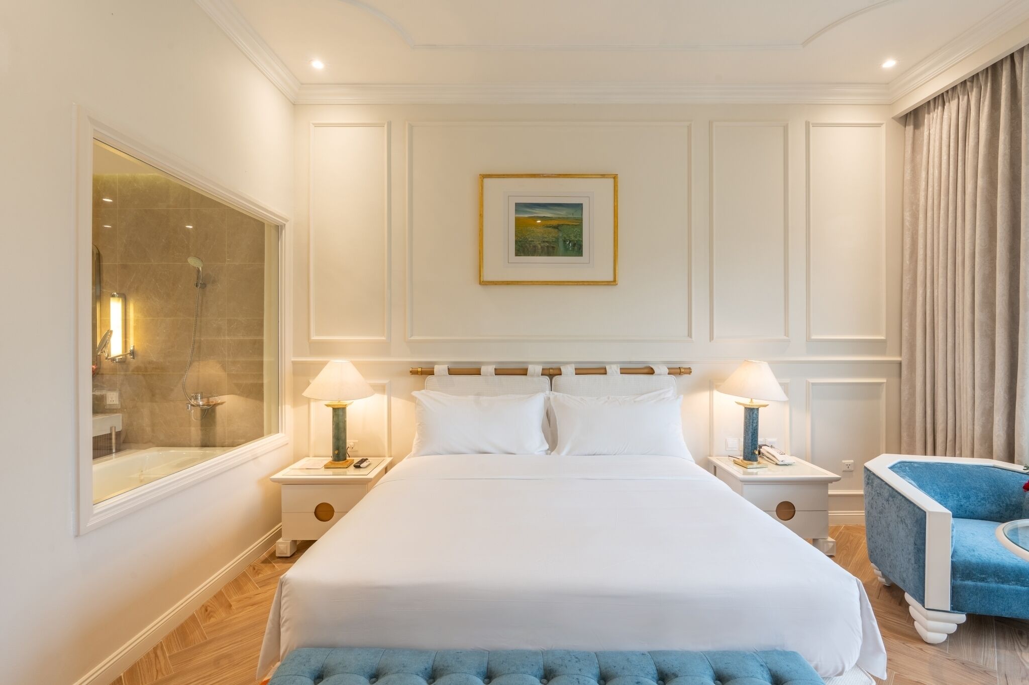 Una habitación de hotel con una cama grande, dos mesitas de noche con lámparas, un ventanal y un sillón azul.