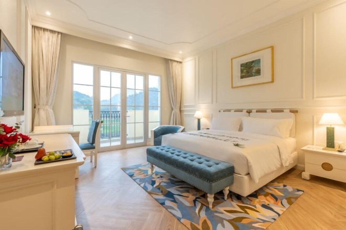 A hotel room with a large bed, a desk with a chair, a small bench, a nightstand, and a window with a view of the outdoors.