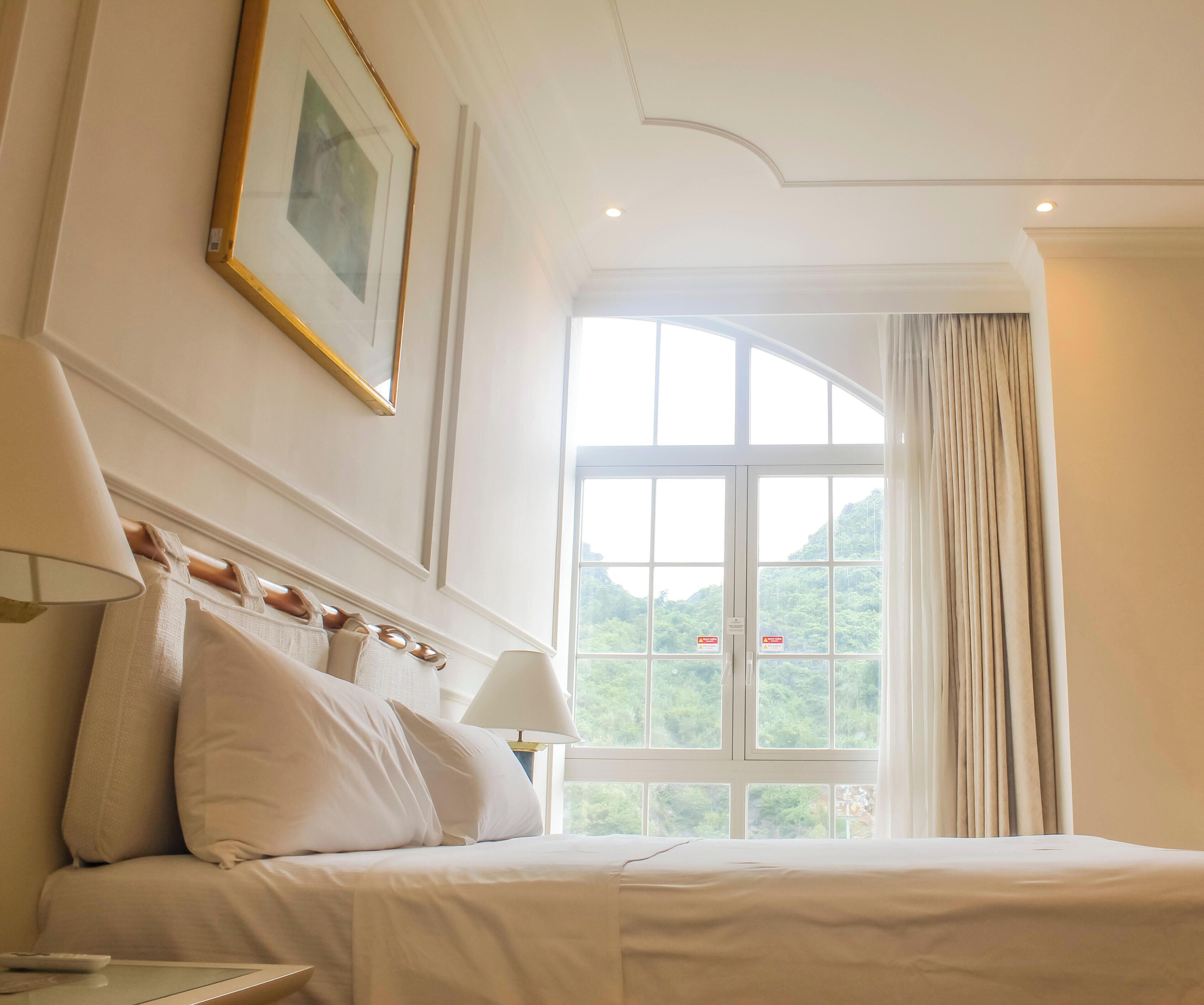 Una habitación de hotel con una cama, un ventanal con cortinas, una lámpara y un cuadro en la pared.