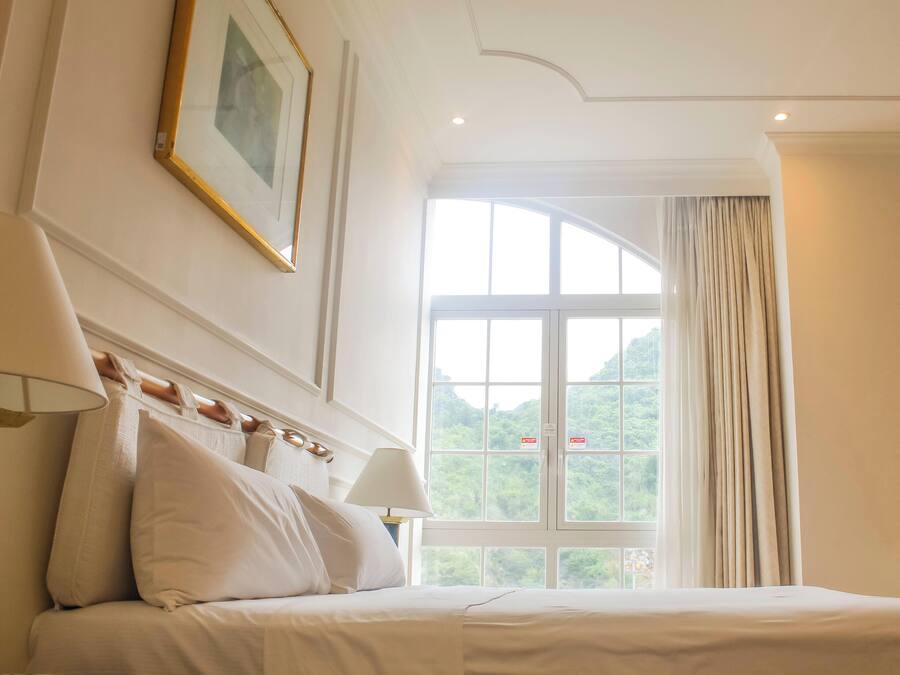 Una habitación de hotel con una cama, un ventanal con cortinas, una lámpara y un cuadro en la pared.
