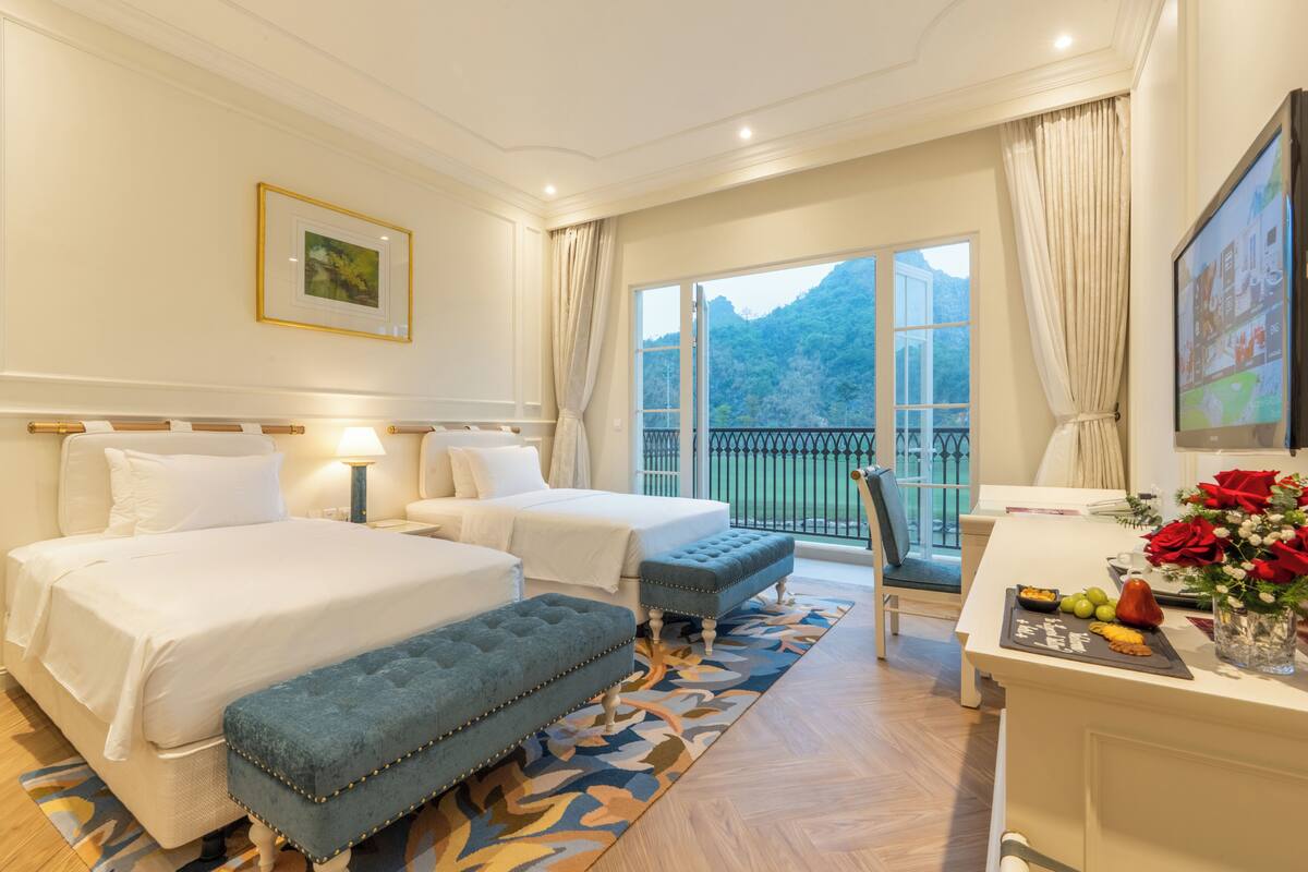 Une chambre d’hôtel avec deux lits, un bureau, une chaise, un balcon offrant une vue sur les montagnes et un coin repas avec une table et des chaises.