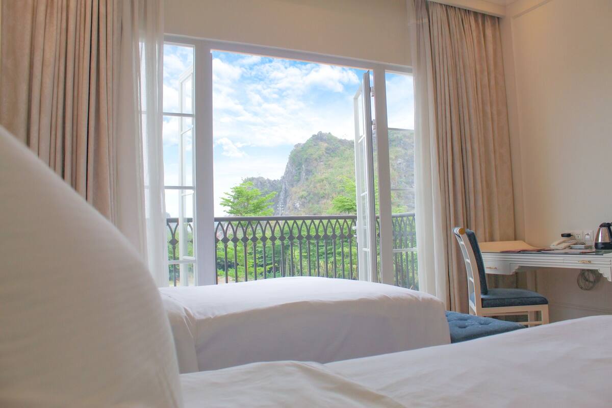 Une chambre d’hôtel avec deux lits, un bureau avec une chaise et un balcon offrant une vue sur un paysage montagneux.