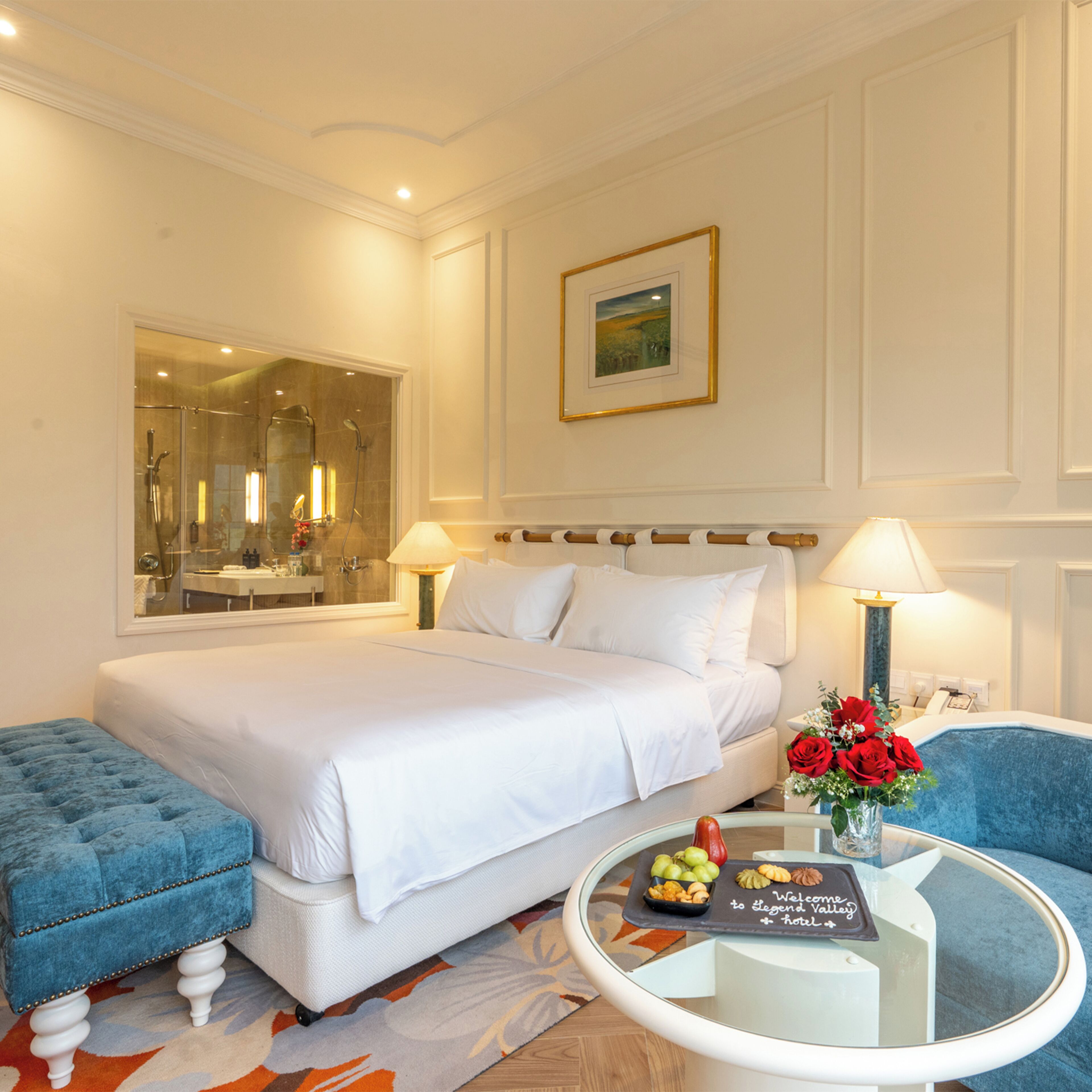 Habitación de hotel con una cama grande, una zona de estar con una mesa de centro, un espejo y un cuadro en la pared.