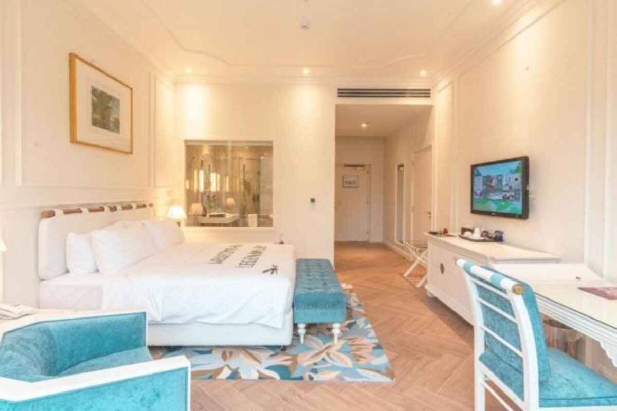 A hotel room with a bed, a desk, a chair, a TV, and a bathroom visible through a glass door.
