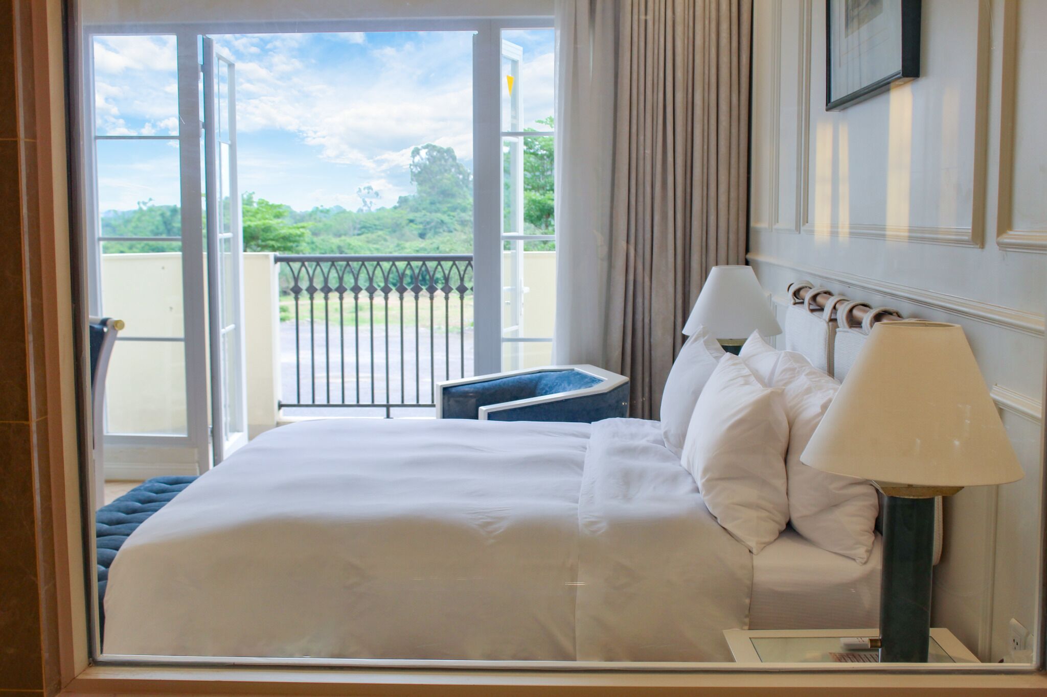 A hotel room with a bed, two bedside lamps, a nightstand, a window with a view of greenery, and a balcony with a railing.