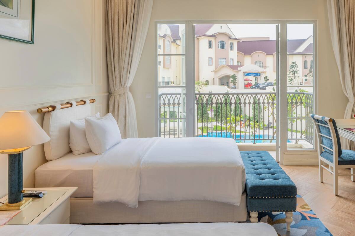 Une chambre d’hôtel avec un lit, une table de chevet avec une lampe, un banc et une vue sur les bâtiments environnants et une piscine.