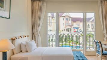 A hotel room with a bed, a bedside table with a lamp, a bench, and a view of the outside buildings and a pool.
