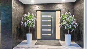 A modern building entrance with a dark brick facade, a central door with horizontal panels, and two potted flowering plants on either side. - 6 bedroom stunning home in Podglavica (Podglavica)