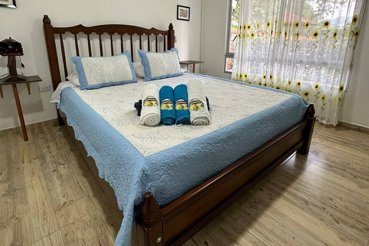 Un lit bien fait, avec une couverture bleue et des oreillers, une tête de lit en bois et une fenêtre ornée de rideaux à motifs floraux.