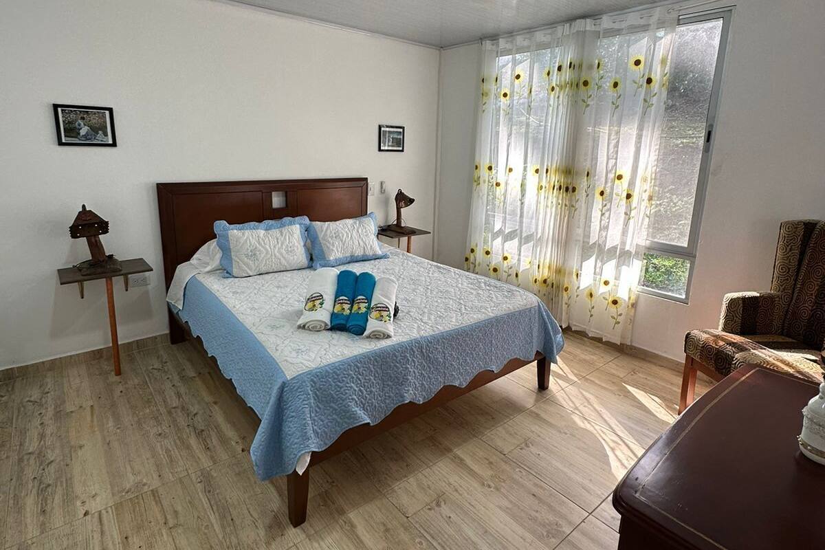 Une chambre à coucher avec un lit en bois, une table de chevet, une lampe, une fenêtre avec des rideaux à motifs floraux et une chaise.